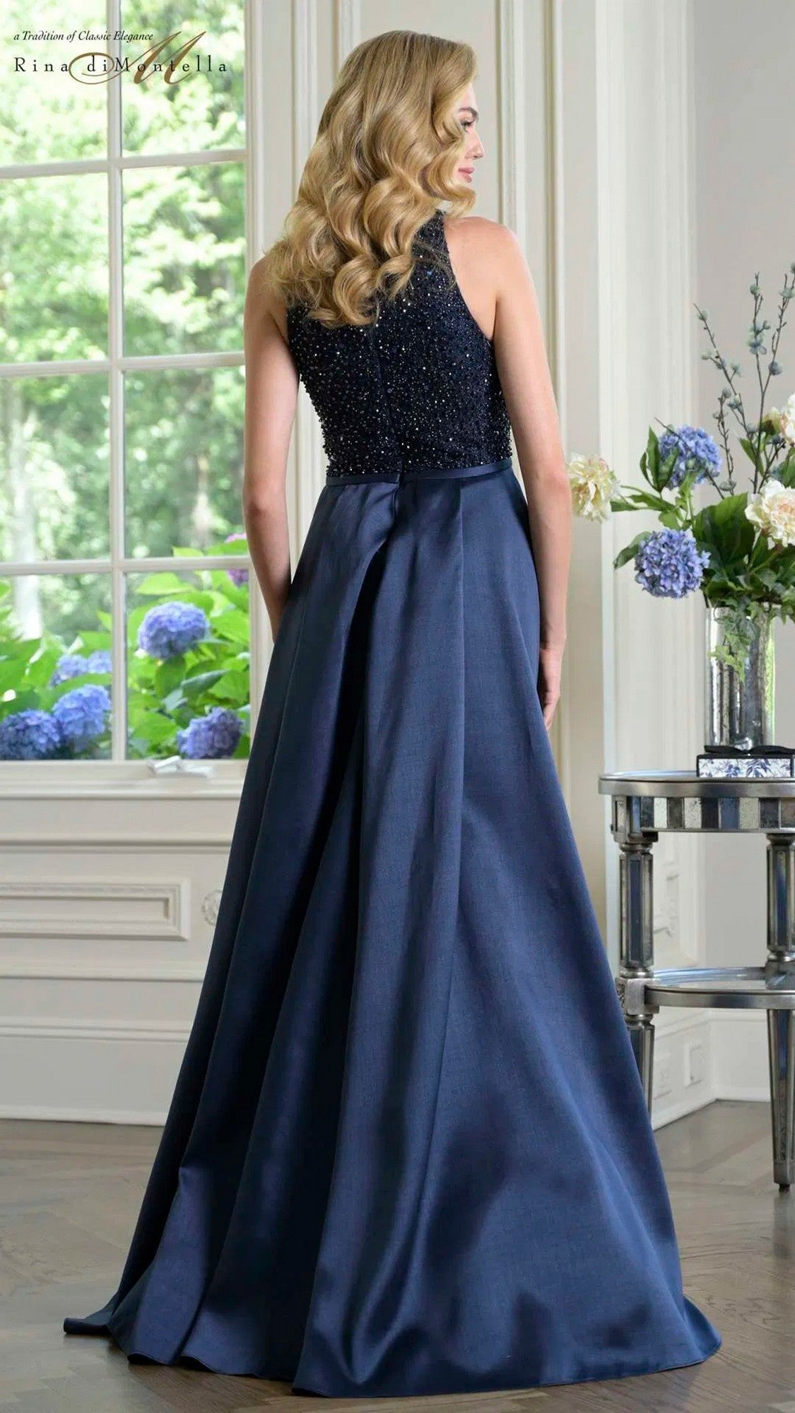 A woman with wavy blonde hair stands indoors in the navy blue Rina Di Montella RD3404 formal dress, featuring a beaded bodice and elegant floor-length skirt, facing away from the camera near a window and a flower-adorned table.